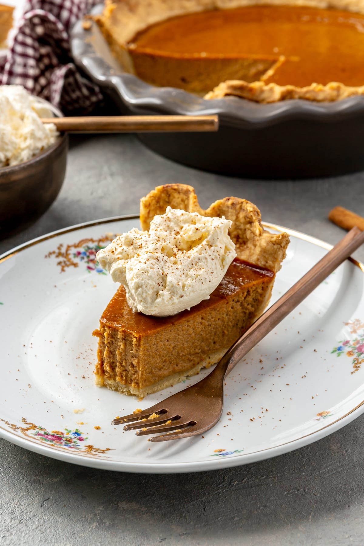 A bite has been taken out of a slice of pumpkin pie, topped with whipped cream and cinnamon. It sits on a white plate with a copper fork. The remaining pie and whipped cream sits in the background.