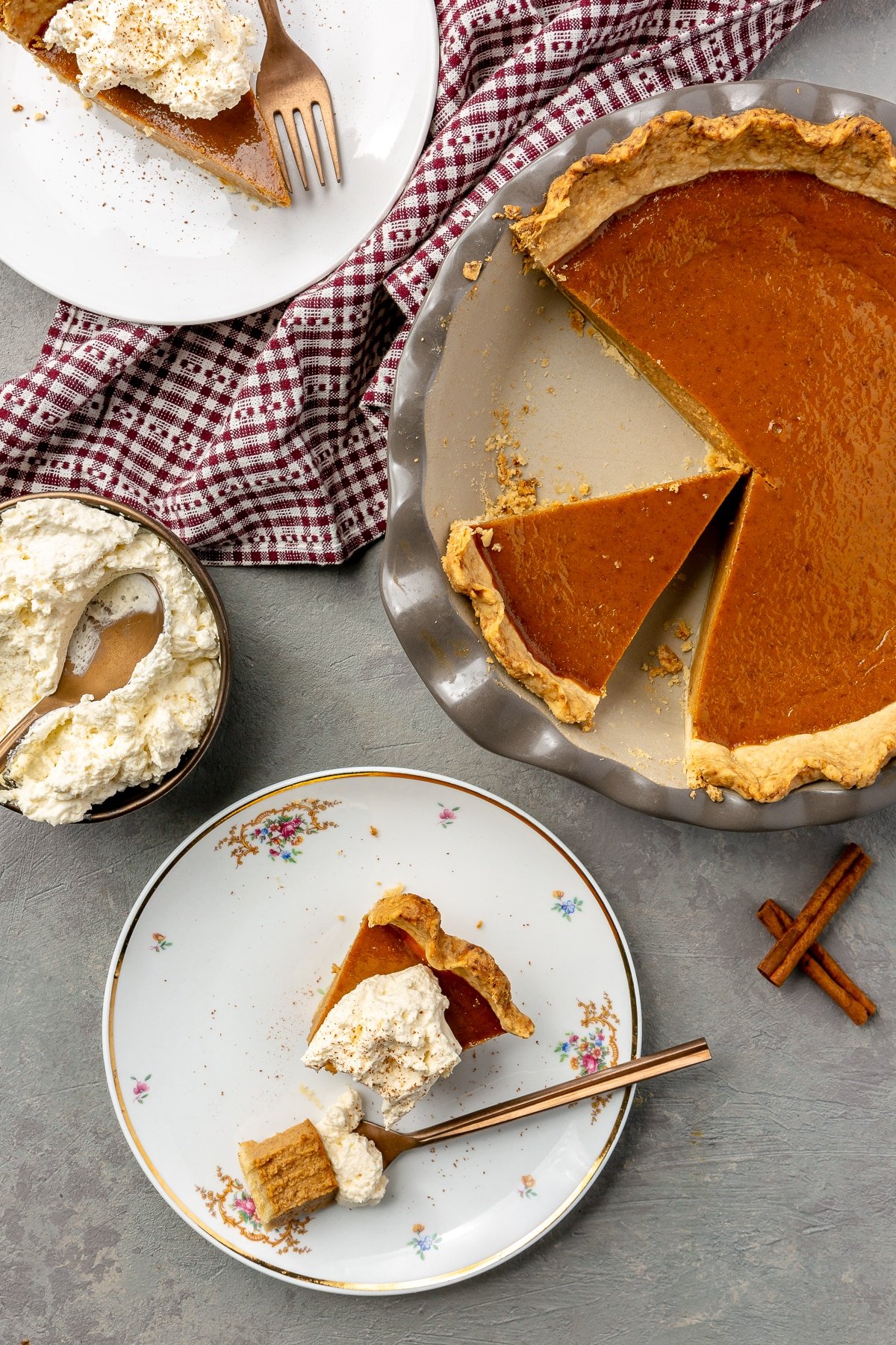 Two slices of pie, one with a bite removed, have been removed and paces on white plates with a copper fork. The remaining pie, whipped cream, and cinnamon sit to the side.