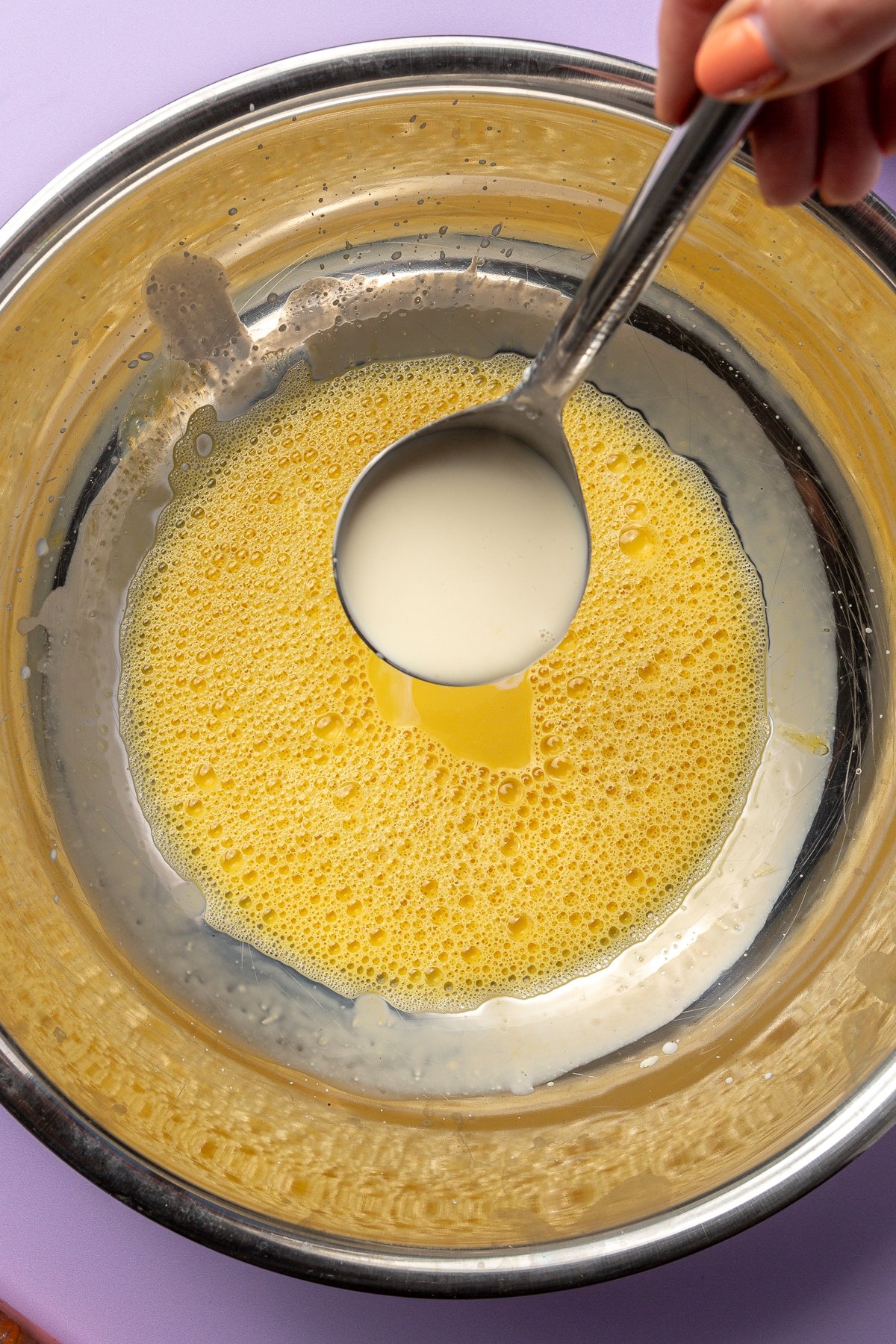 A hand is shown spooning in small amounts of the milky mixture into the egg mixture.