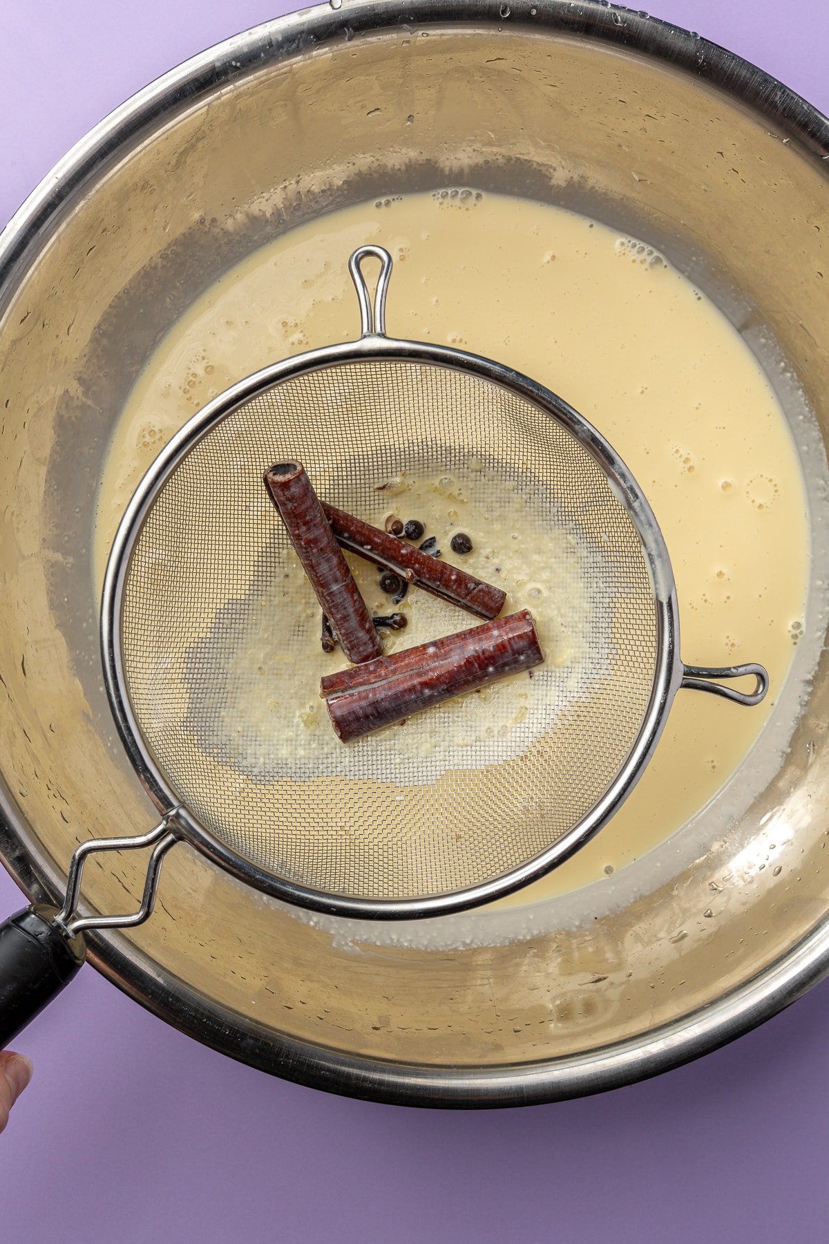 A colander is shown scooping out cinnamon sticks and cloves from the milky mixture.
