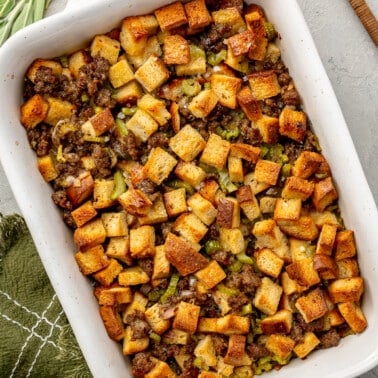 Fully baked stuffing sits in a white enameled, rectangular, baking dish. A metal serving spoon, green dish cloth, and several sprigs of rosemary and sage, sit to the side.