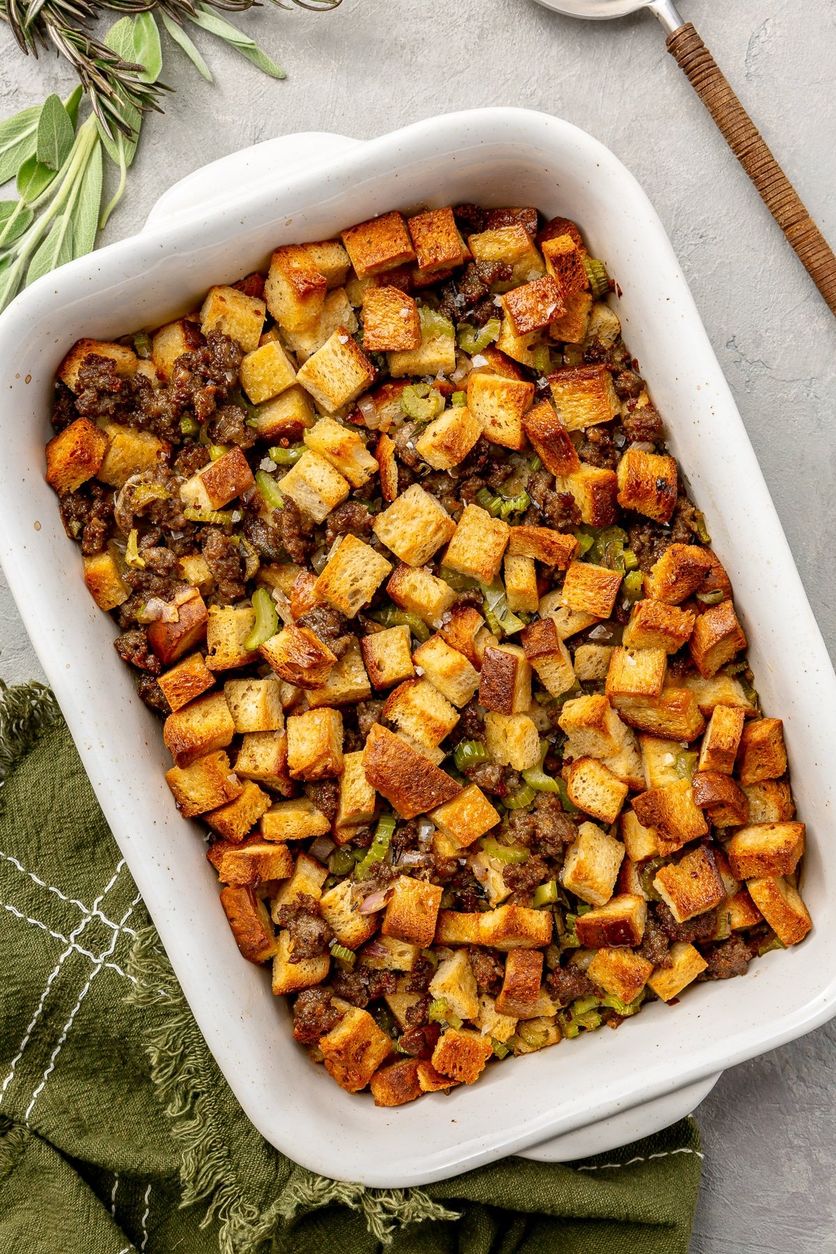 Fully baked stuffing sits in a white enameled, rectangular, baking dish. A metal serving spoon, green dish cloth, and several sprigs of rosemary and sage, sit to the side.