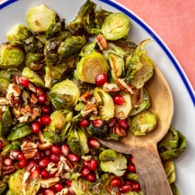 The brussels sprouts have been placed into a white, oval-shaped serving dish with a blue border. Pomegranate seeds and chopped pecans have been added on top. A wooden serving spoon is shown scooping the brussels sprouts.