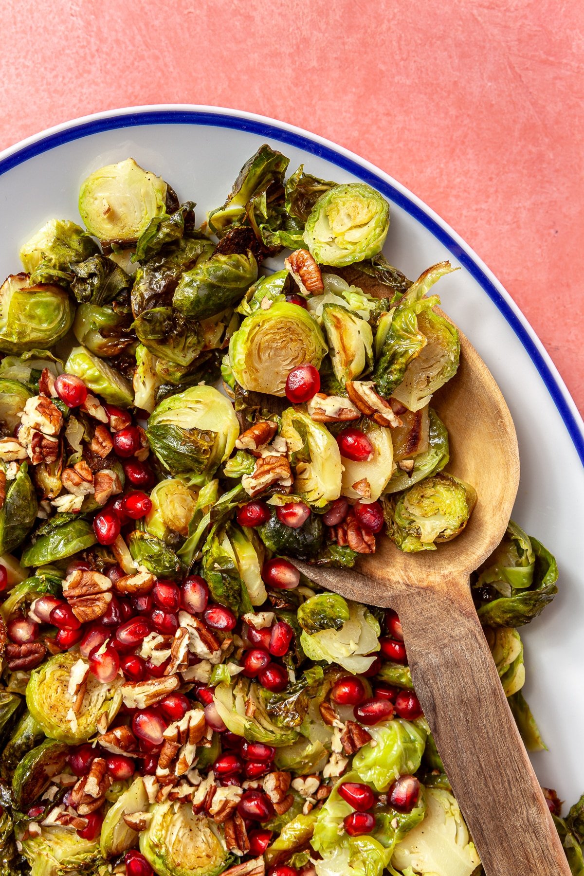 The brussels sprouts have been placed into a white, oval-shaped serving dish with a blue border. Pomegranate seeds and chopped pecans have been added on top. A wooden serving spoon is shown scooping the brussels sprouts.