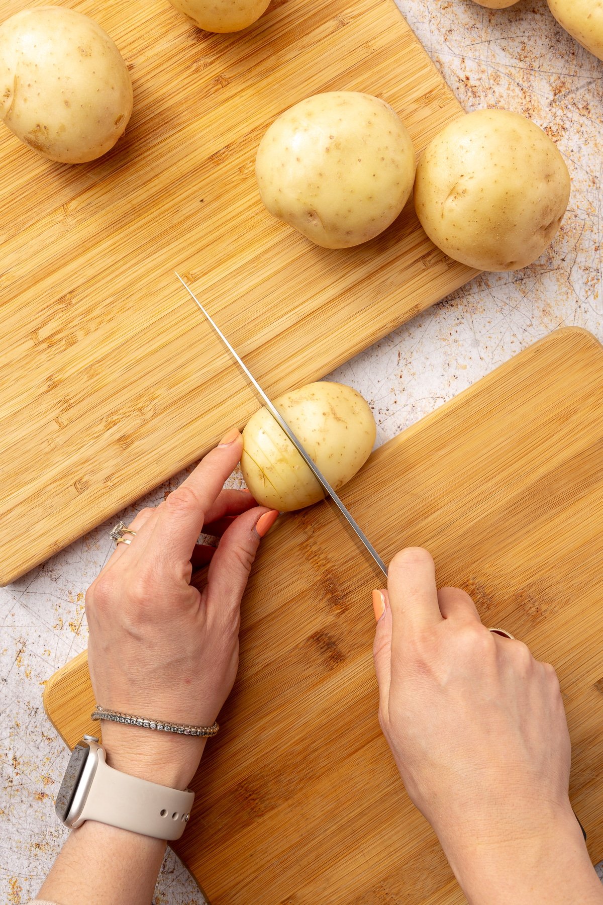 Two hands are shown cutting the potato which sits in between the two cutting boards. The reming potatoes sit to the side.