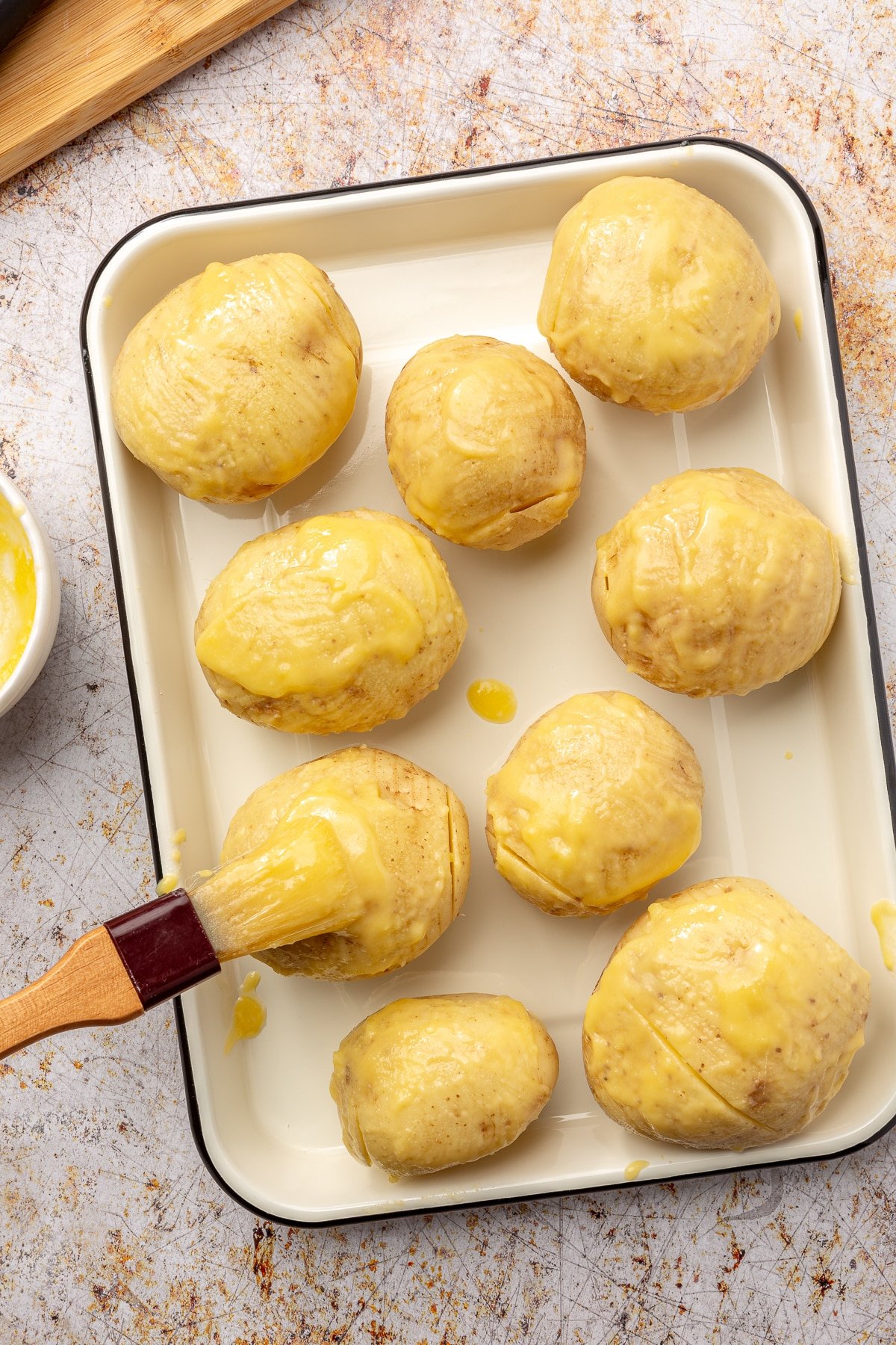 Sliced potatoes sit on a baking pan. A brush is shown dressing them in a thick yellow sauce.