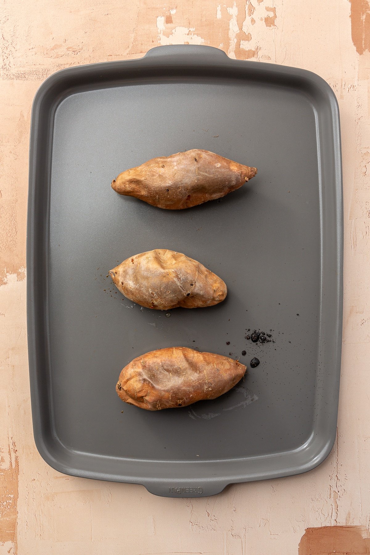 Three, now baked, sweet potatoes sit on a grey baking pan.