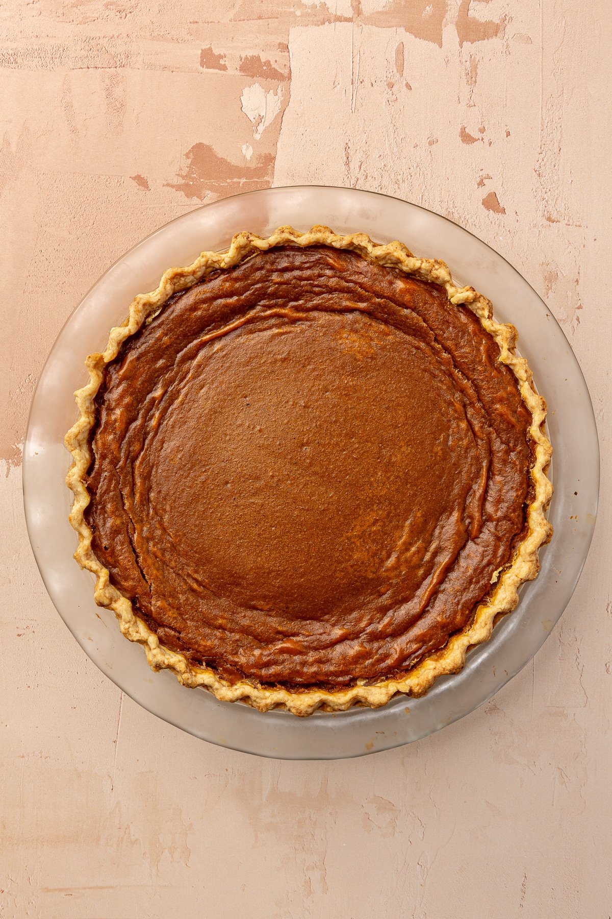 A fully cooked, now dark brown tin color, sweet potato pie remains in its glass pie dish on a muted pink background.