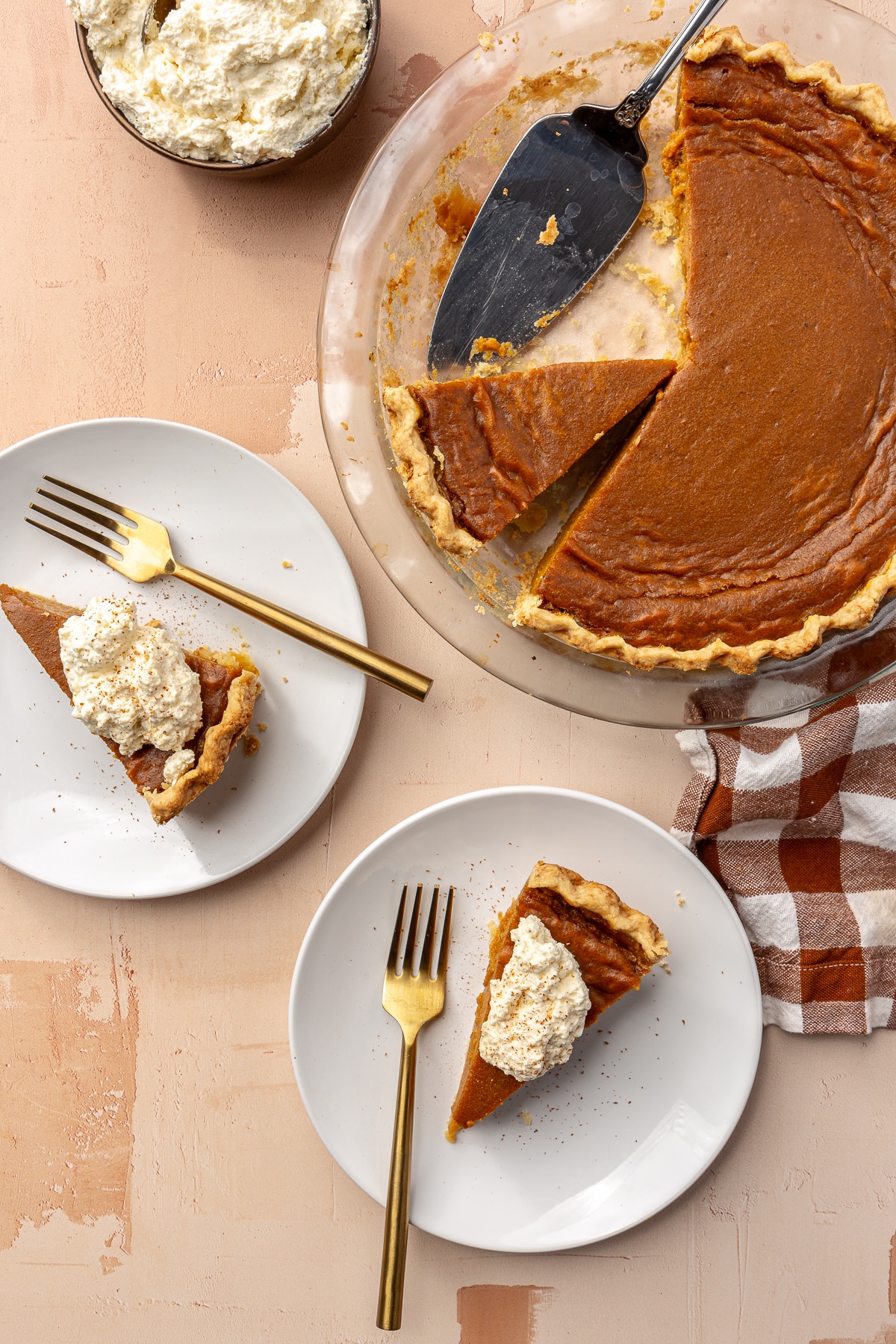 Three slices. of sweet potato pie have been served on white plates with golden forks. The third piece and the remaining pie sits in a glass pie dish. A bowl of whipped cream sits to the side.