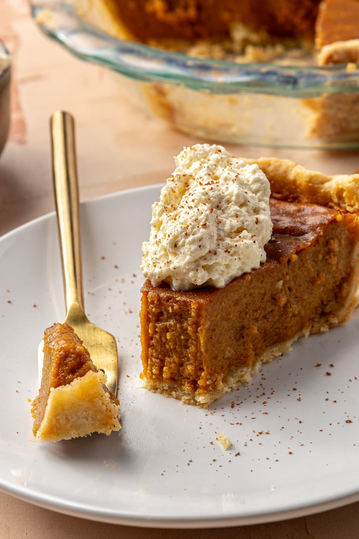 A slice of sweet potato pie, topped with whipped cream and ground nutmeg, sits on a white plate. A bite has been taken out and sits on a golden fork.