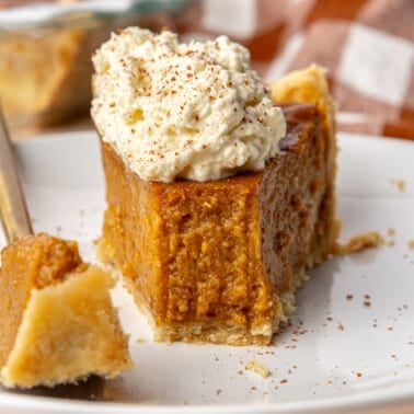 A slice of sweet potato pie, topped with whipped cream and ground nutmeg, sits on a white plate. A bite has been taken out and sits on a golden fork.