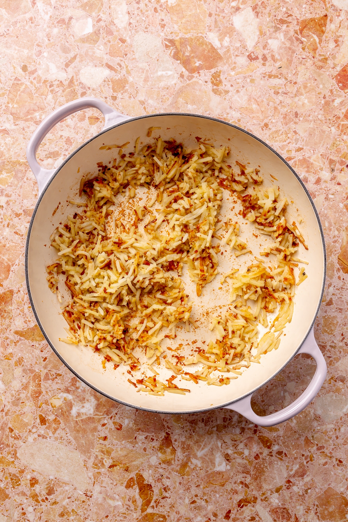 Lightly browned hash browns sit in a white enameled pot which sits on a pink countertop.