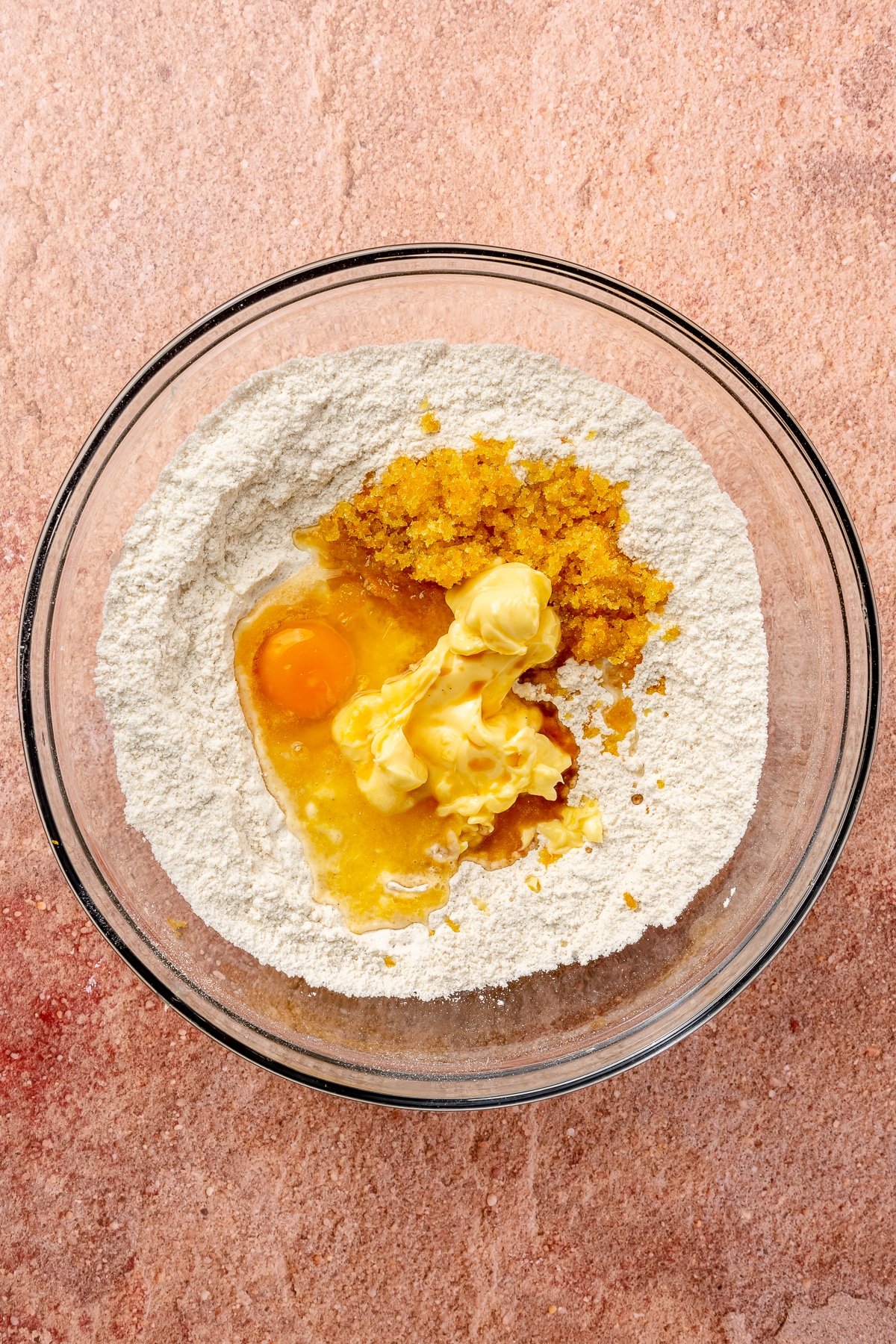 Flour, an egg, butter, vanilla, and the orange sugar mixture sit in a large glass mixing bowl. It sits on a light pink countertop.