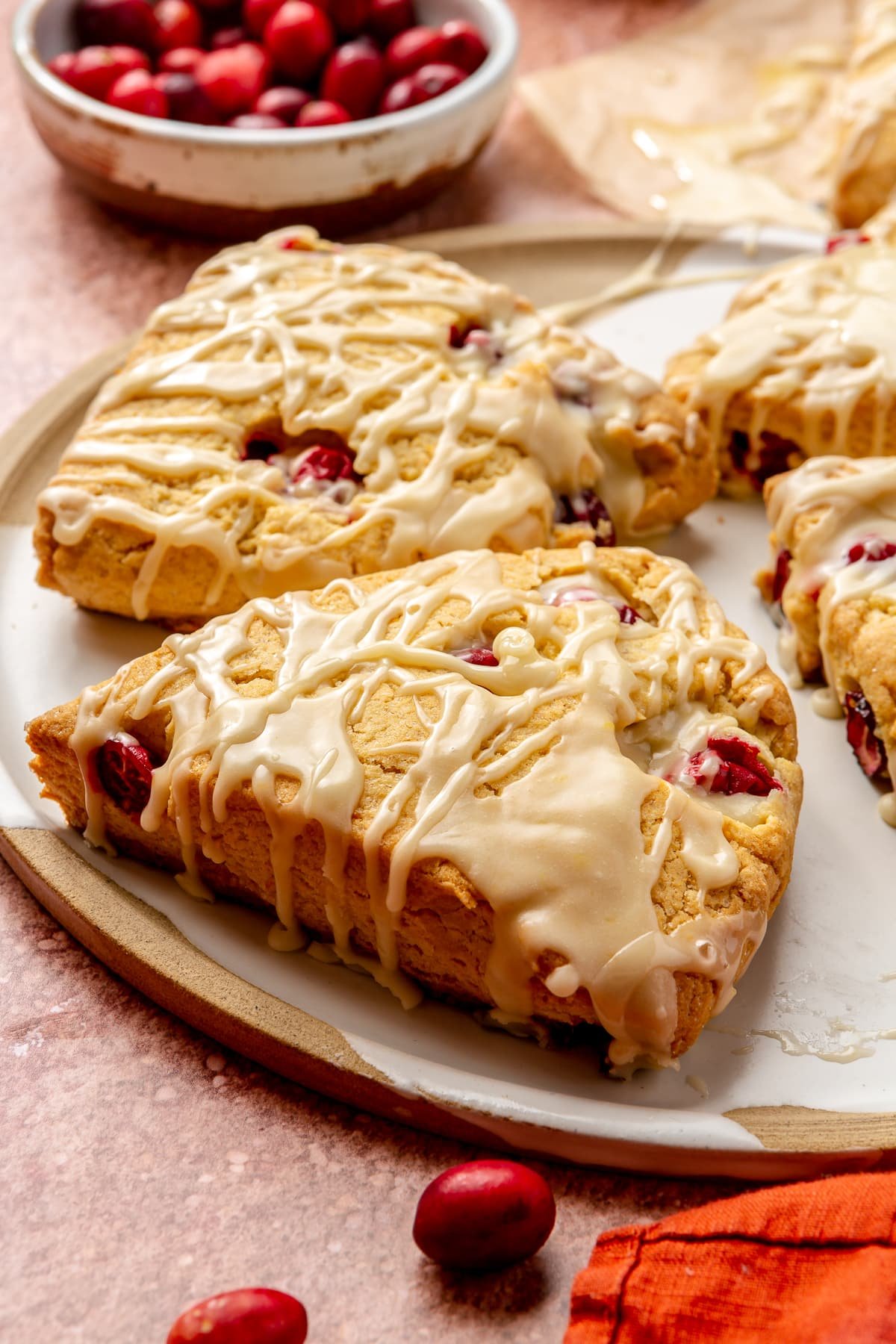 Four cranberry orange scones sit on a brown and white serving plate. A red dish cloth and small bowl of cranberries sit to the side.