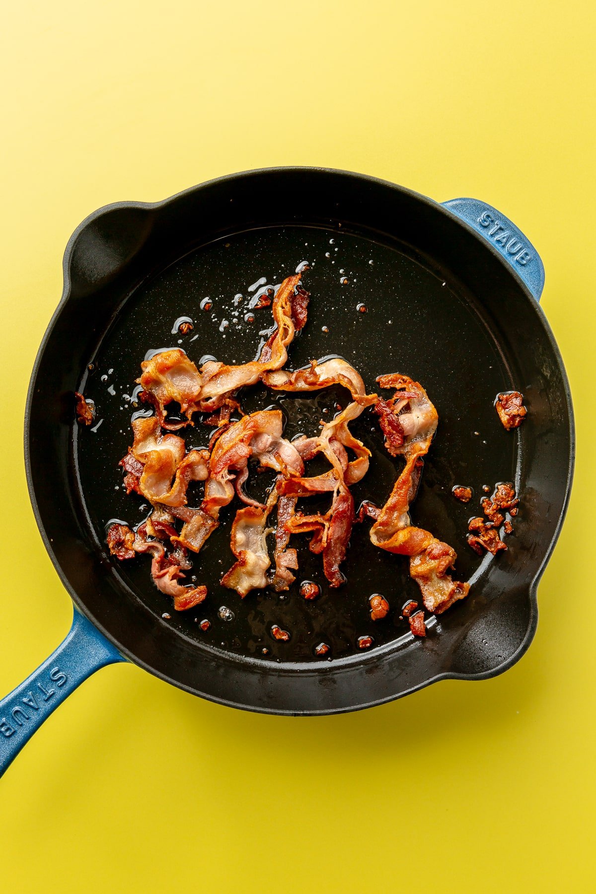 Strips of bacon are shown being cooked in a black cast-iron skillet with blue handles.
