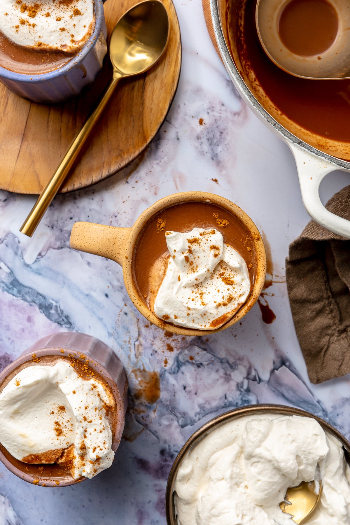 Three mugs of Mexican hot chocolate, each with a dollop of whipped cream and sprinkle of cinnamon, sit on a marble countertop. A gold spoon sits to the side along with a brown dishcloth. The remaining hot chocolate, in the white pot, sits in the upper right-hand corner.