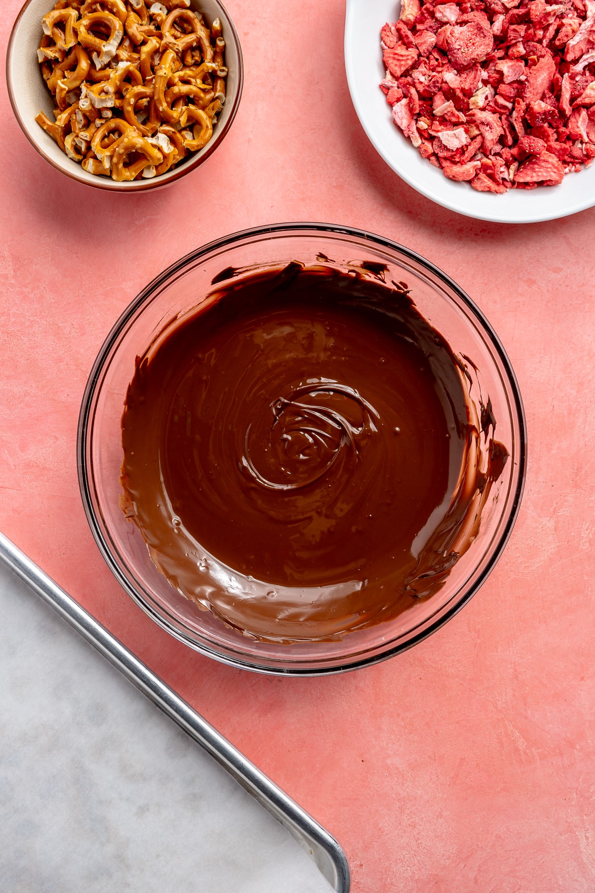 Melted chocolate sits in a glass bowl. A bowl of pretzels and a bowl of freeze-dried strawberries sit to the side.