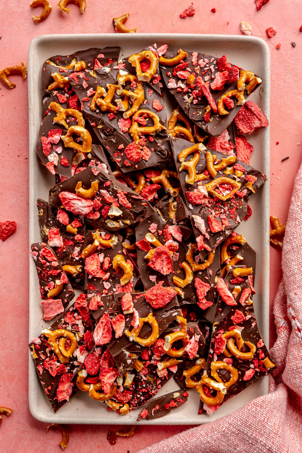 Dark chocolate bark with strawberries and pretzels sits on a light grey serving tray which sits on a light pink countertop. A pink dish cloth sits to the side.