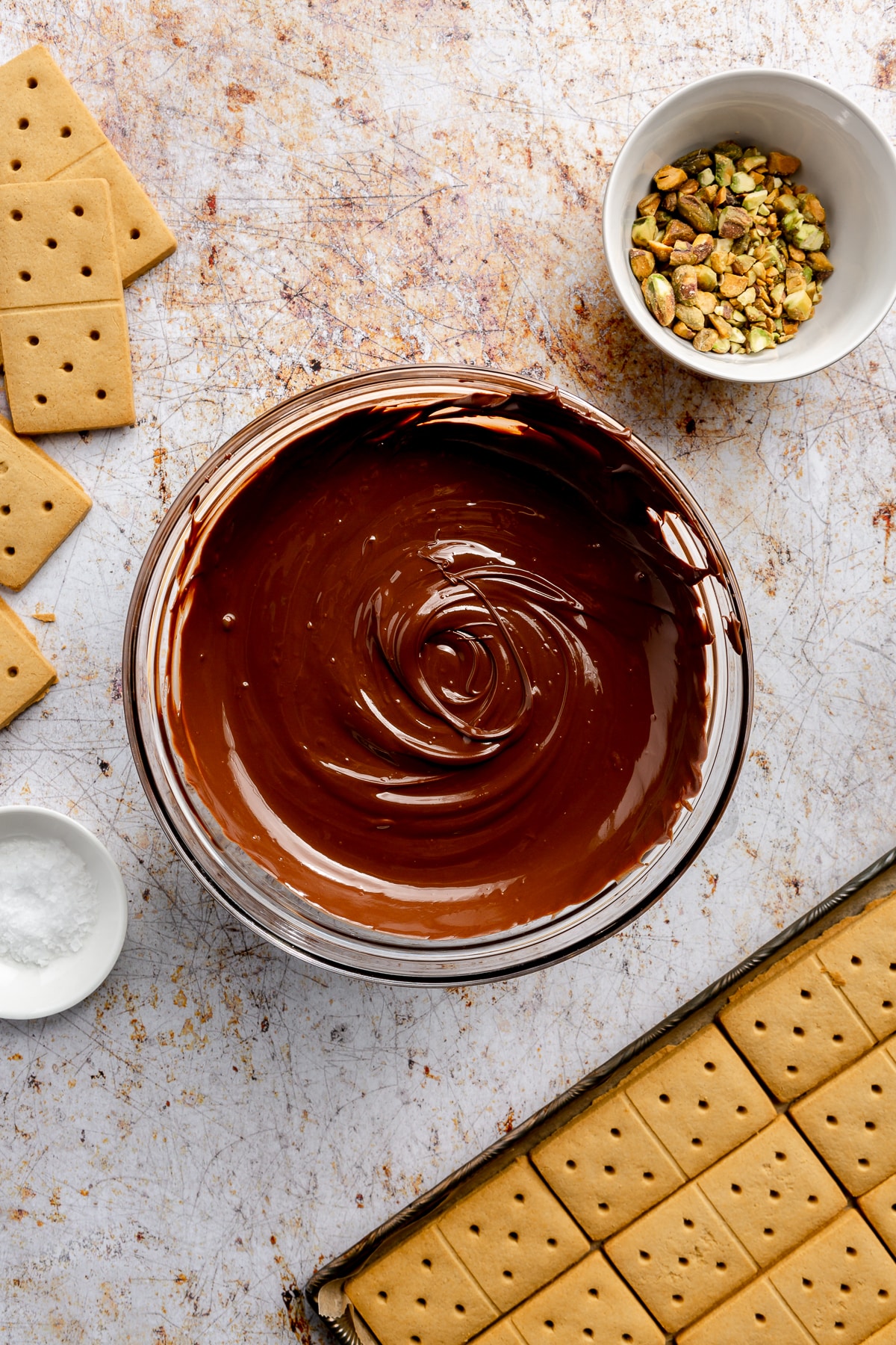 Melted chocolate sits in a glass bowl. The tray of graham crackers and a bowl of pistachios sit to the side.
