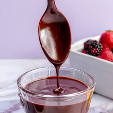 Ganache sits in a glass cup on a marble countertop. A metal spoon is shown lifting up some of the ganache to show consistency.