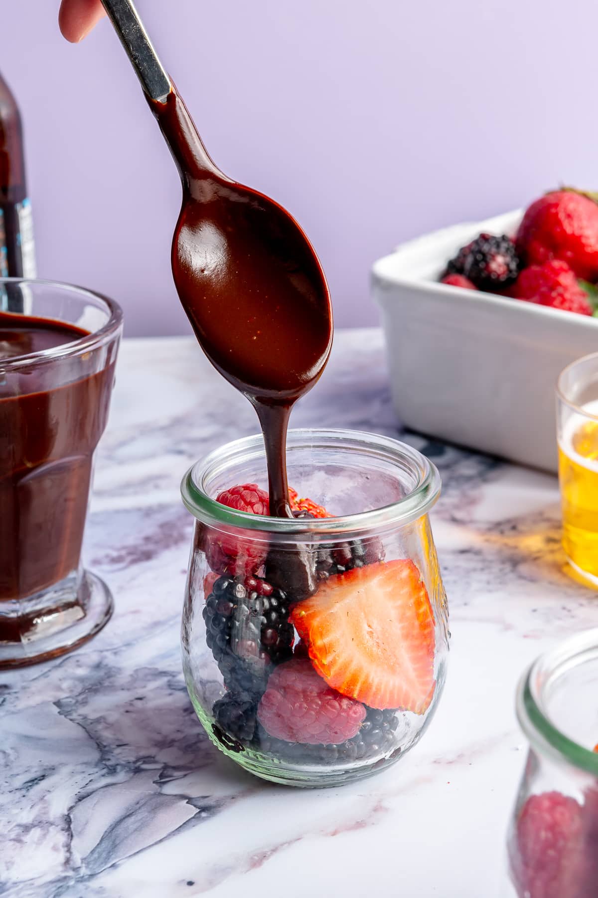 A metal spoon is shown drizzling ganache over a glass cup of fresh berries.