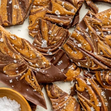 Salted chocolate freezer bark sits on a white tray. A small wooden bowl of flaky sea salt sits to the side.