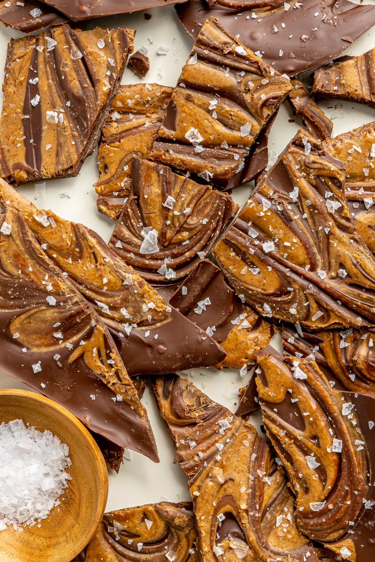 Salted chocolate freezer bark sits on a white tray. A small wooden bowl of flaky sea salt sits to the side.
