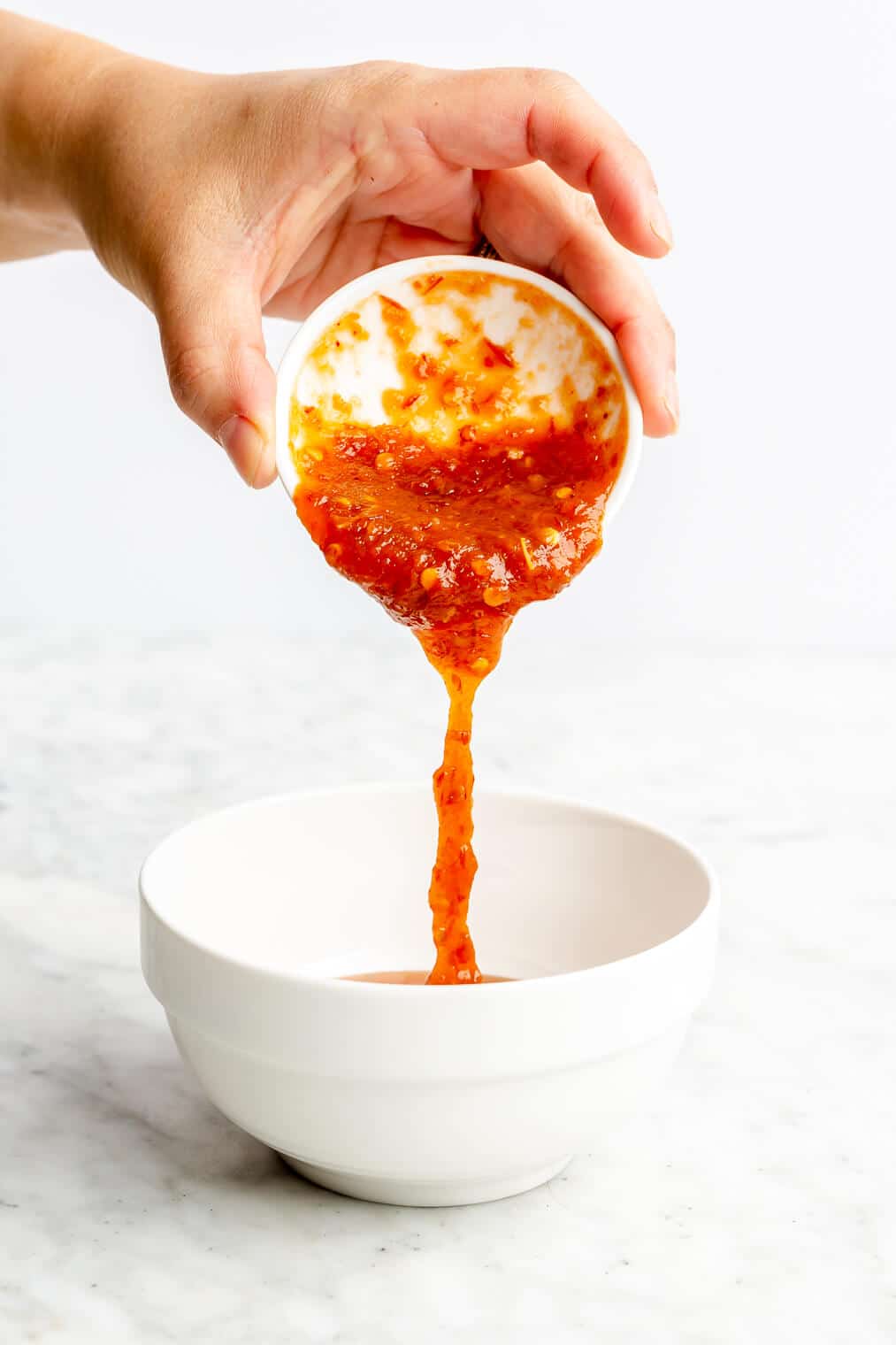 A person pouring chili garlic sauce into a small white bowl.