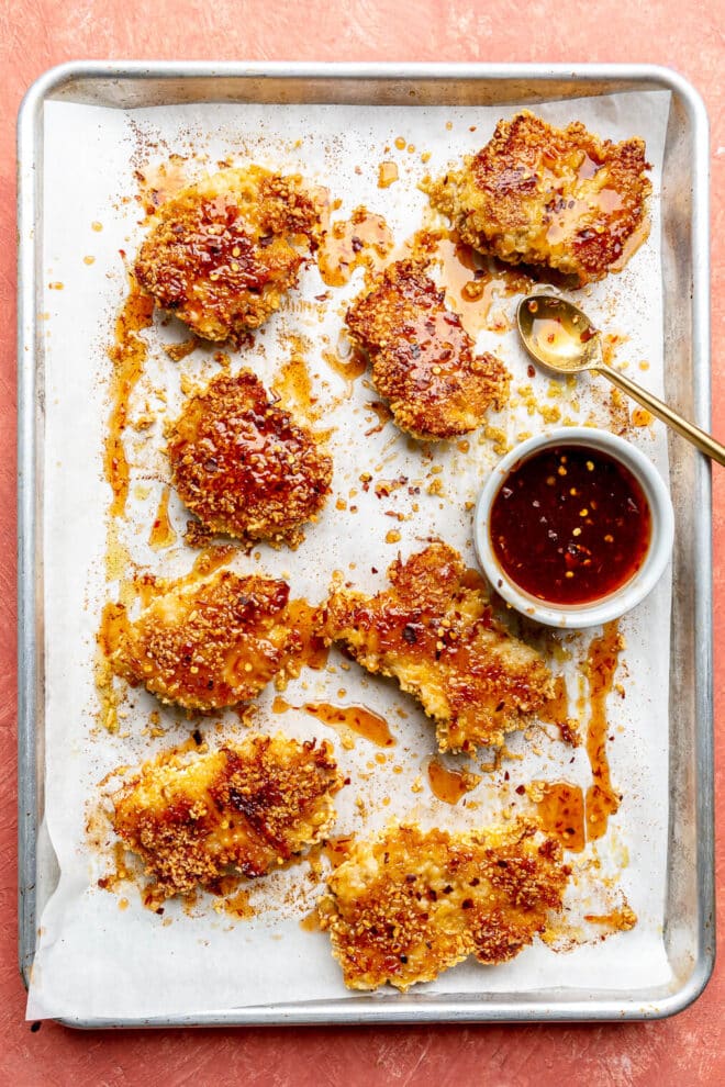 Several hot honey chicken thighs on a parchment paper lined sheet pan next to a bowl of hot honey sauce.