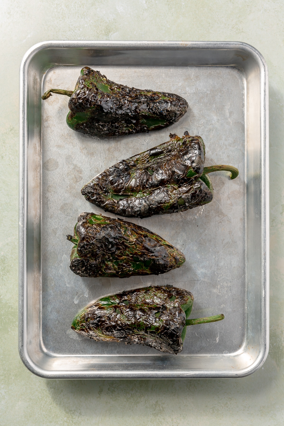 4 charred and blackened green peppers sit on a metal tray.