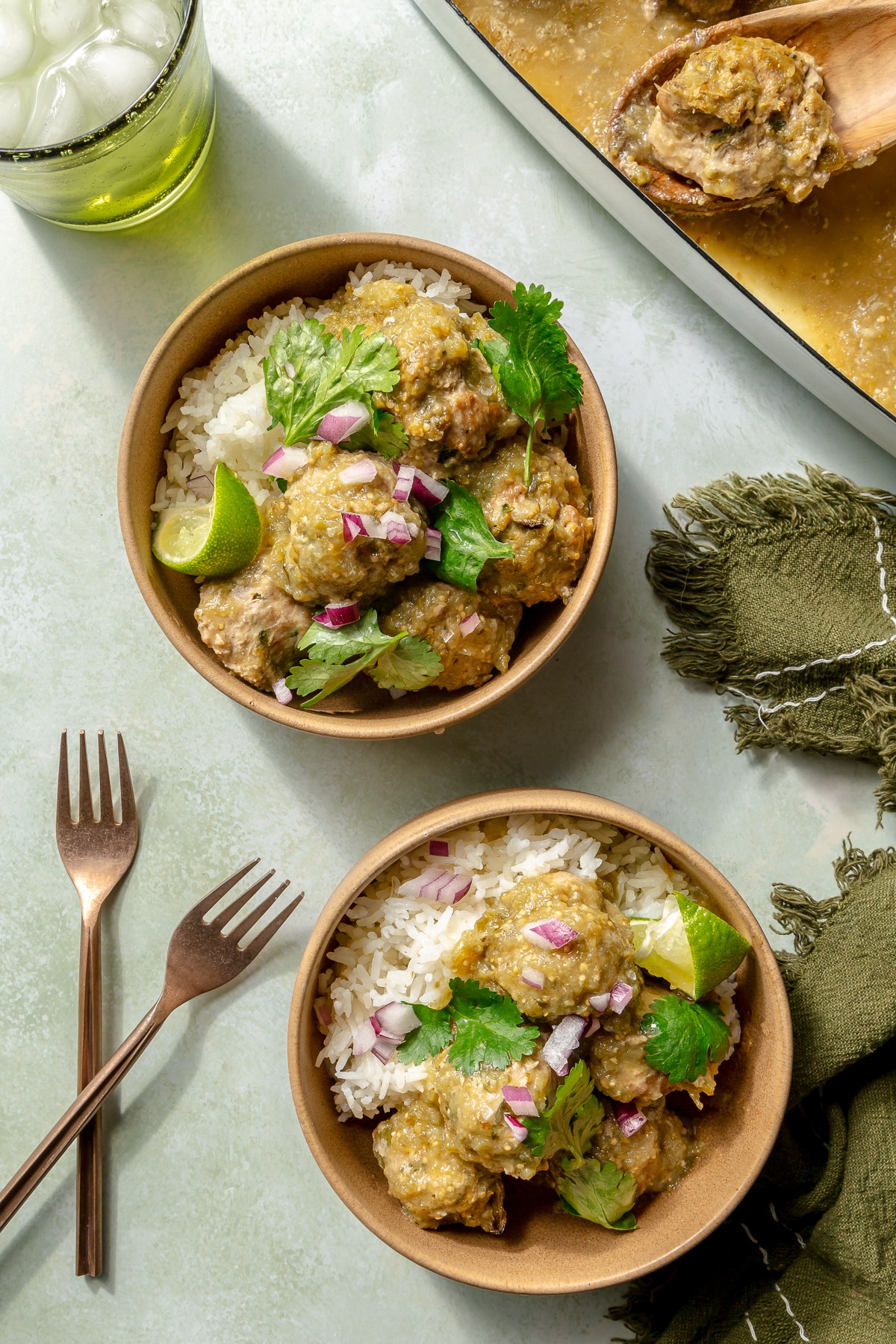 Enchilada verde turkey meatballs have been served in two bowls over white rice. Diced onions and cilantro have been placed on top for garnish. Two copper forks sit to the side.