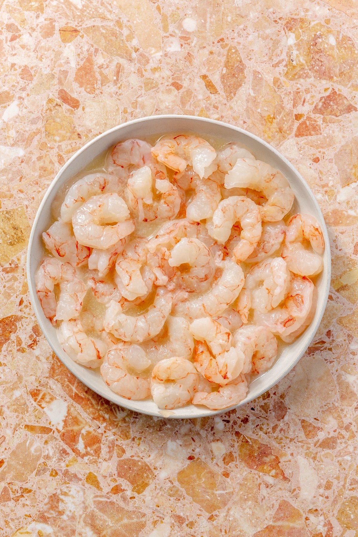 Deveined shrimp sit in a white bowl on a light pink countertop.