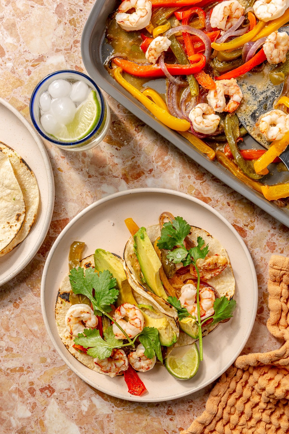 Shrimp fajitas sit on a cream colored plate. The remaining roasted veggies and shrimp sit on the baking tray which sits to the side. An iced beverage sits in the corner.