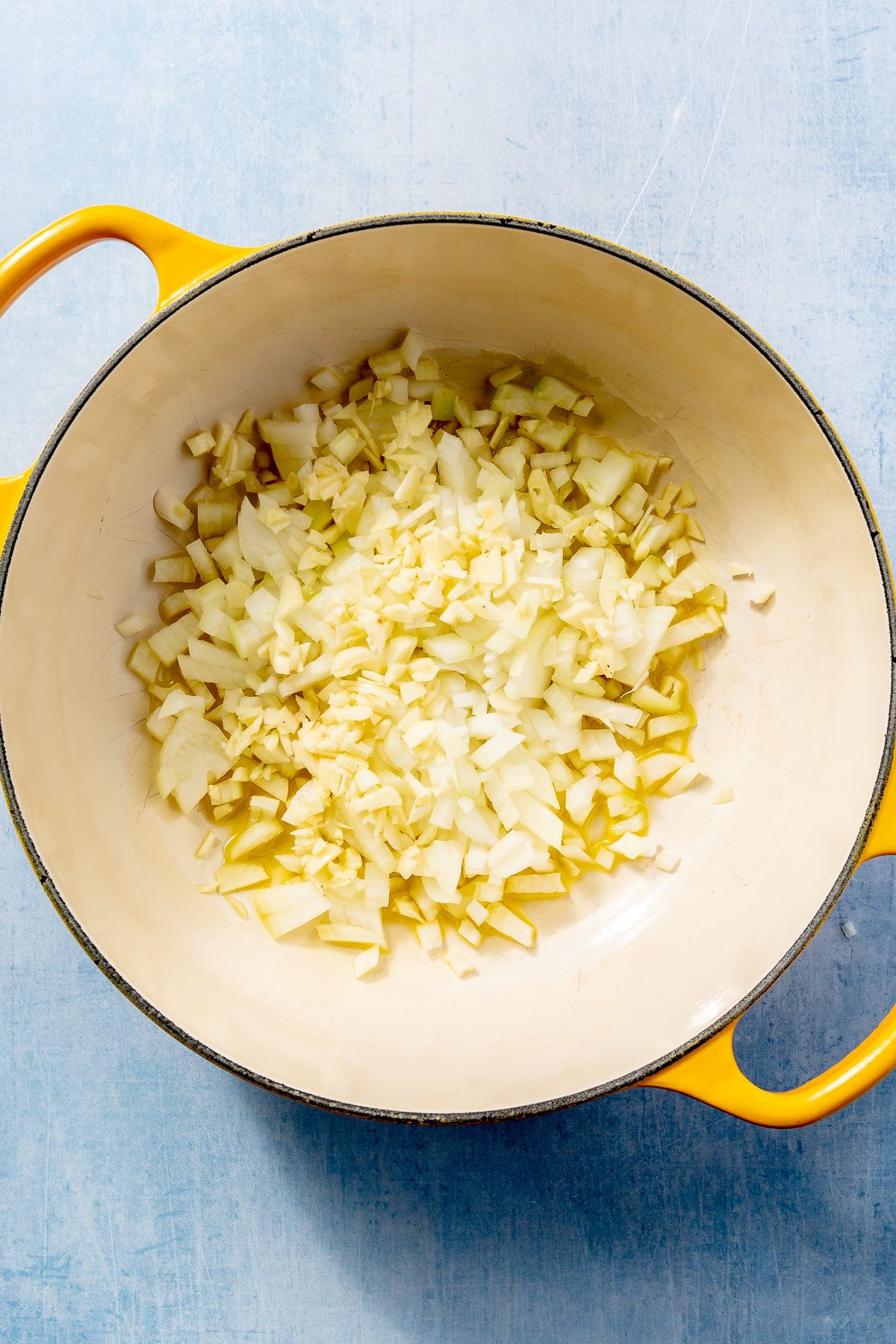 Diced onions, drizzled in oil, sit in a white enameled pot with yellow handles.