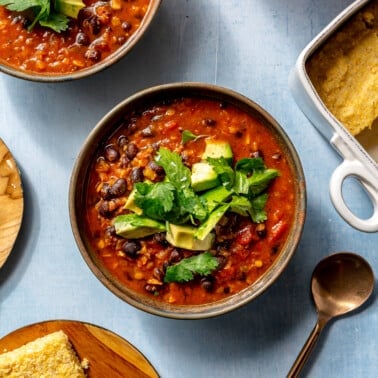 A single serving of chili sits in a brown bowl on a blue countertop. It has been garnished with cilantro and diced avocado. A plate of cornbread and a copper spoon sit to the side.