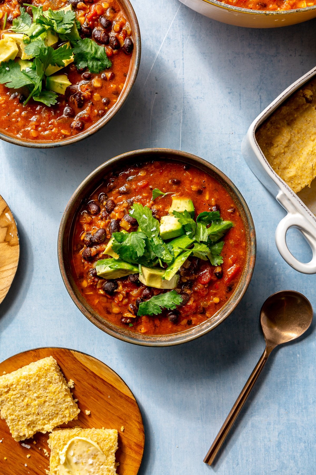 A single serving of chili sits in a brown bowl on a blue countertop. It has been garnished with cilantro and diced avocado. A plate of cornbread and a copper spoon sit to the side.
