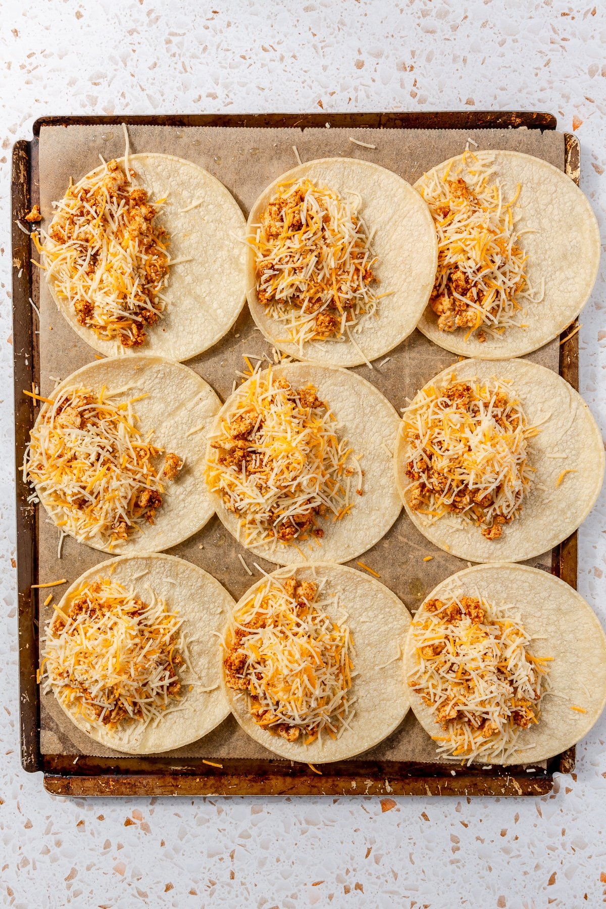 Seasoned ground chicken and shredded cheese have been put on 9 tortillas. They sit on metal baking sheet.
