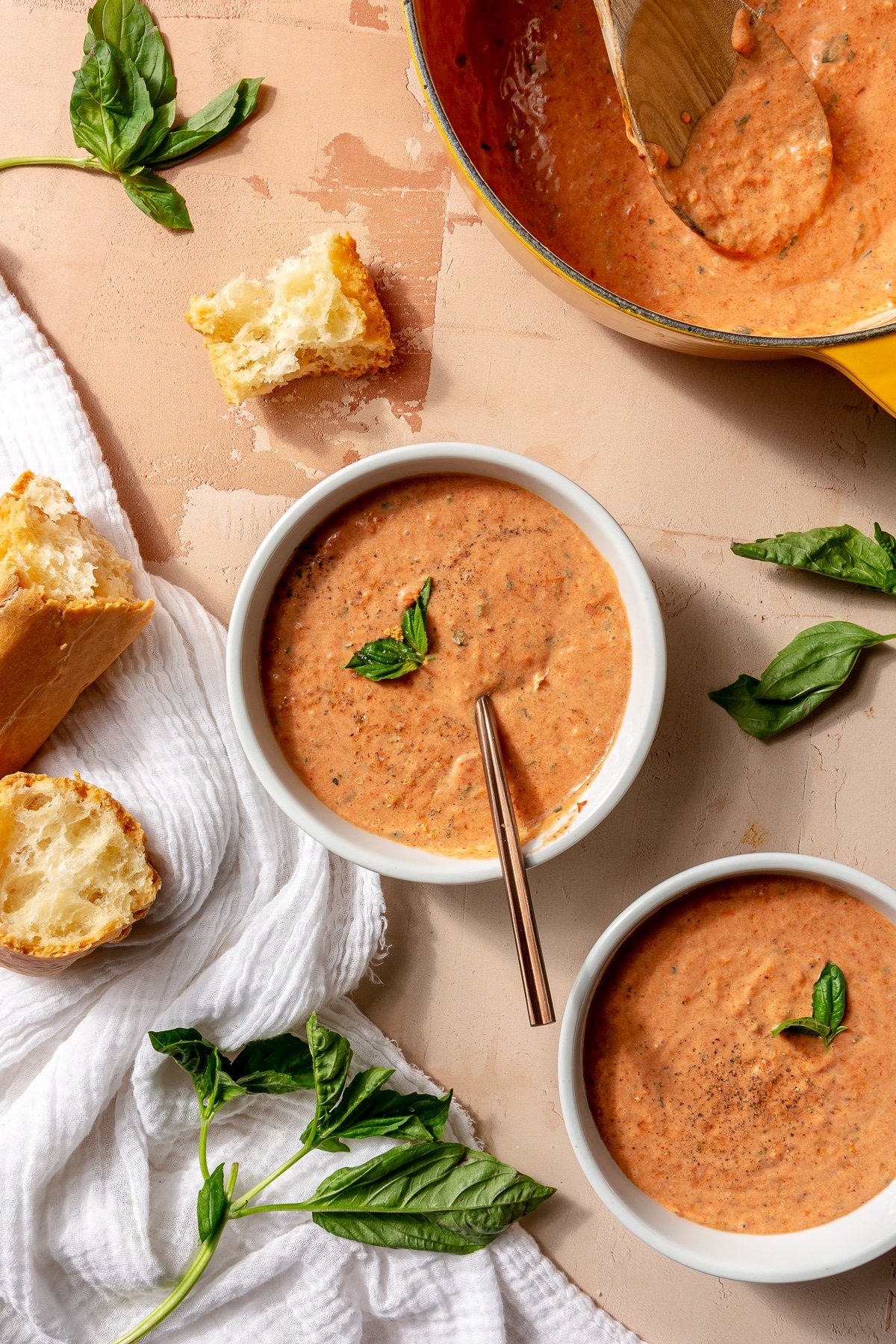 Tomato soup has been served in two white bowls. They each have been toped with basil. A toasted baguette sits to the side.