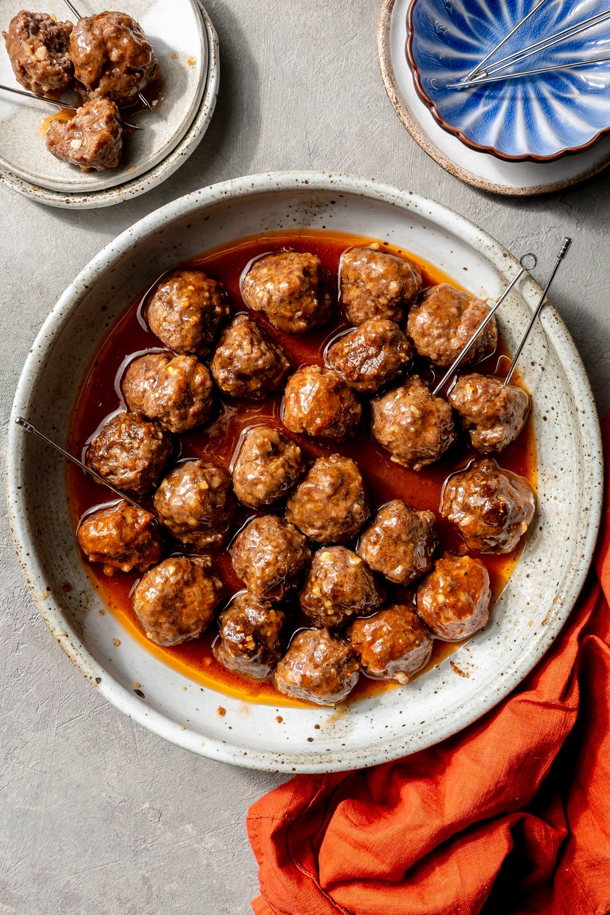 Sweet and sour meatballs have been placed in a serving bowl. Serving skewers have been placed in a couple. A blue serving bowl and red kitchen cloth sit to the side.