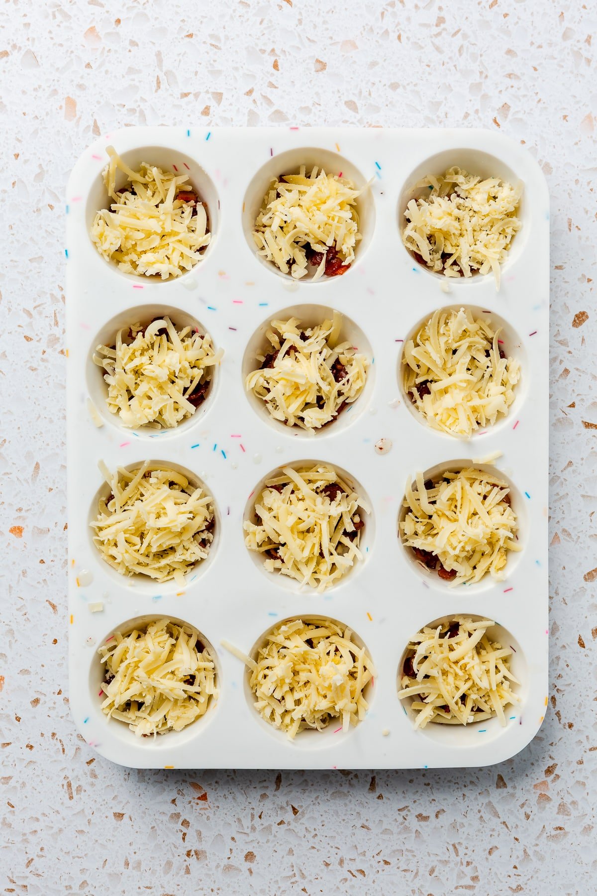 A white shredded cheesed other ingredients have been placed in the bottom of a white, silicone muffin tray.