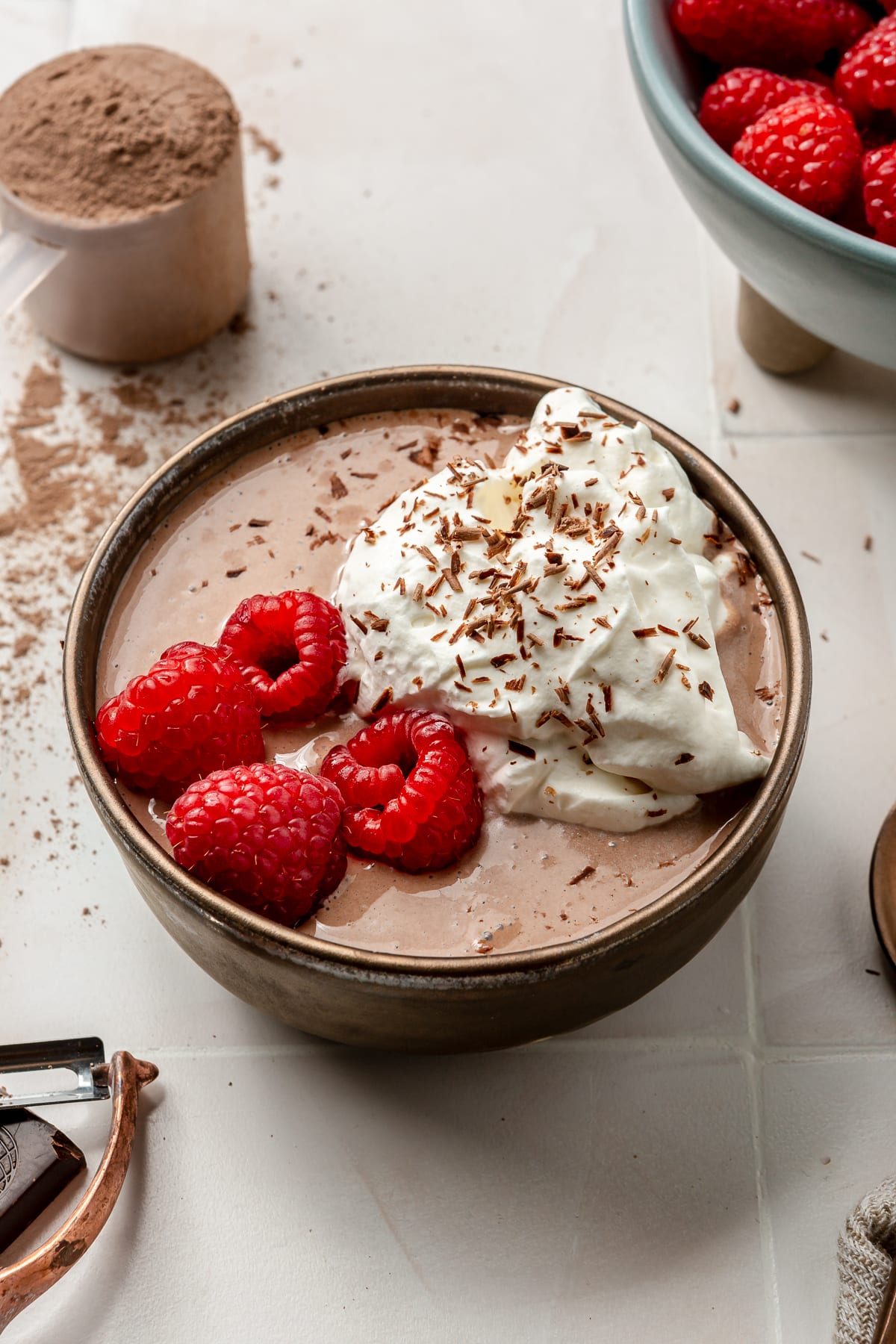 A bowl of chocolate protein pudding sits on a white tiled countertop. It has been topped with raspberries, whipped cream, and chocolate shavings. The remaining raspberries sit in a blue bowl off to the side.