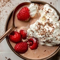 A bowl of chocolate protein pudding sits on a white tiled countertop. It has been topped with raspberries, whipped cream, and chocolate shavings. A copper colored spoon is shown scooping bite.