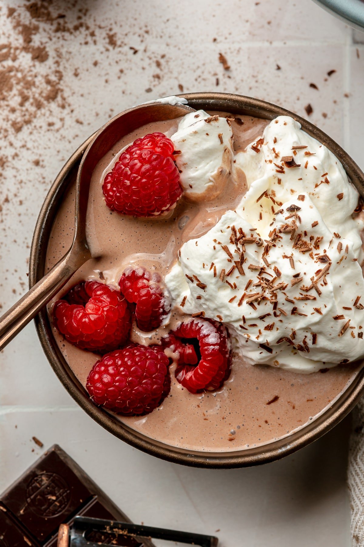 A bowl of chocolate protein pudding sits on a white tiled countertop. It has been topped with raspberries, whipped cream, and chocolate shavings. A copper colored spoon is shown scooping bite.