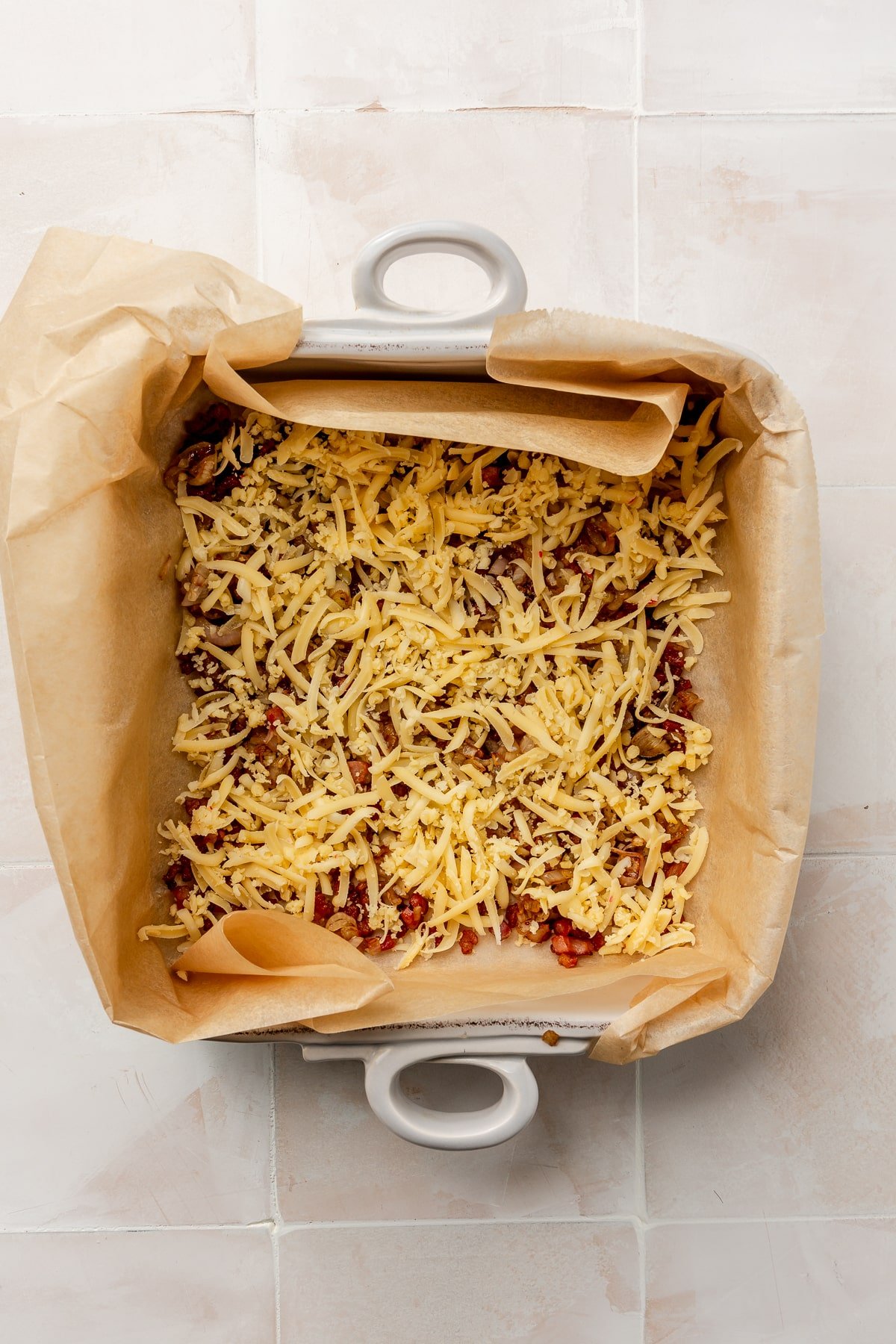 A white shredded cheese and bacon bits have been added to the bottom of a parchment paper lined baking dish.