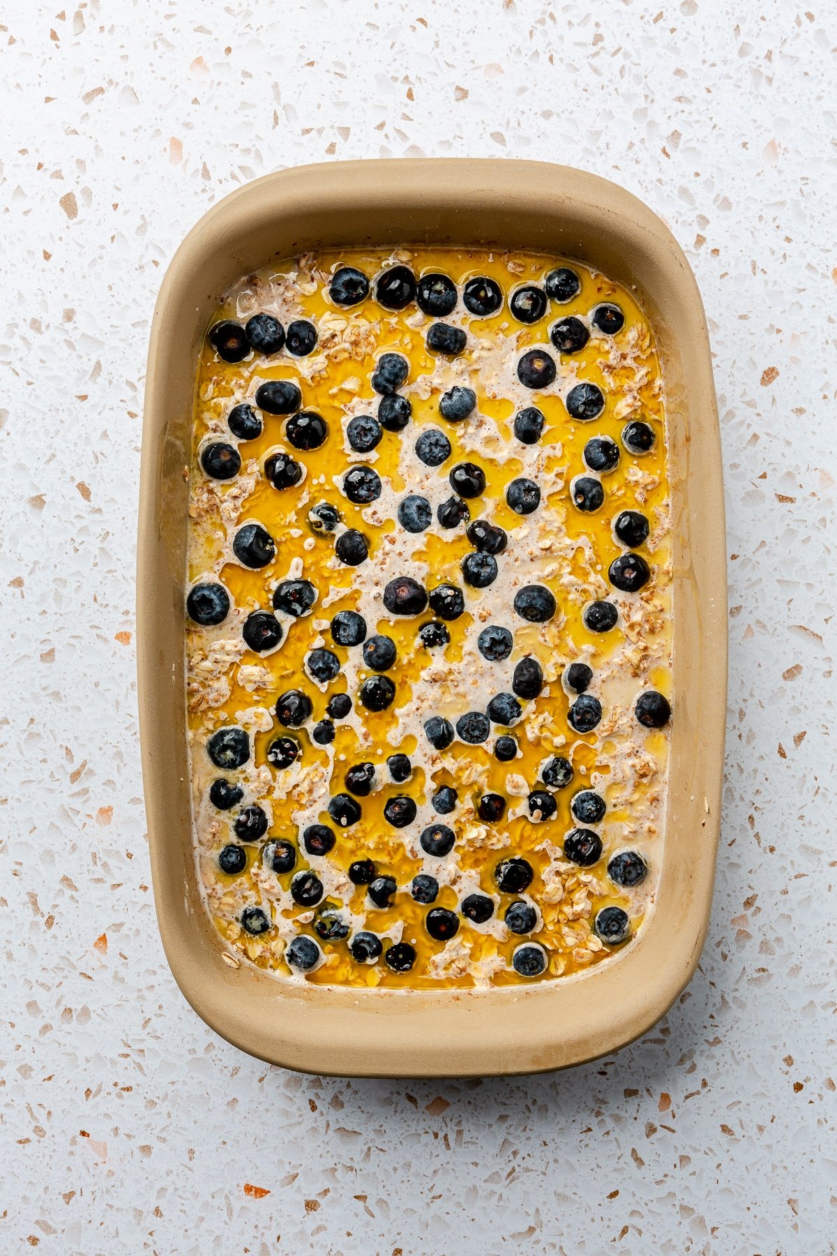 The liquid ingredients have been added over the top of the dry in the beige-colored baking dish. It has been topped with blueberries.