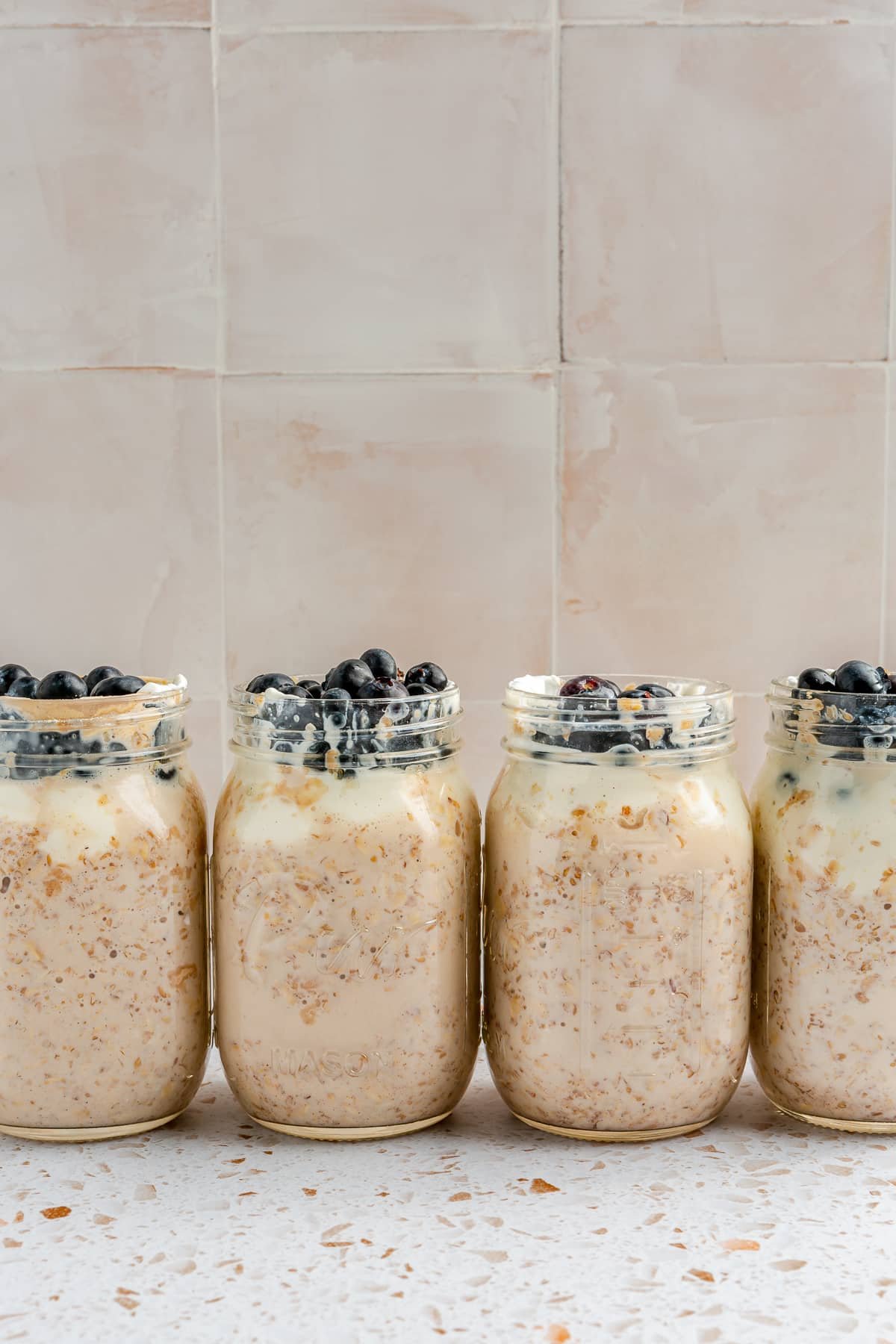 4 mason jars, full of high protein overnight oats, have been topped with blueberries. They sit arranged in a line on a white speckled countertop.