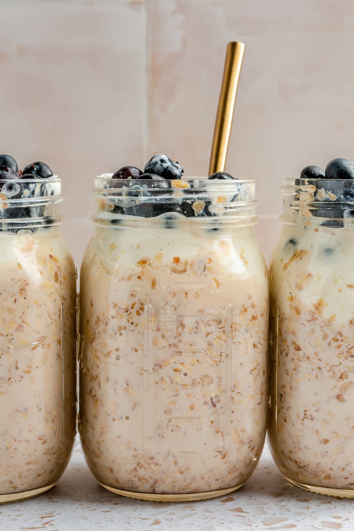 3 mason jars, full of high protein overnight oats, have been topped with blueberries. They sit arranged in a line on a white speckled countertop. A gold-colored spoon has been added to one of the jars.