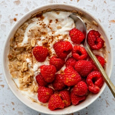 A bowl of hot protein overnight oats sits in a white serving bowl. It has been topped with raspberries and a metal spoon.