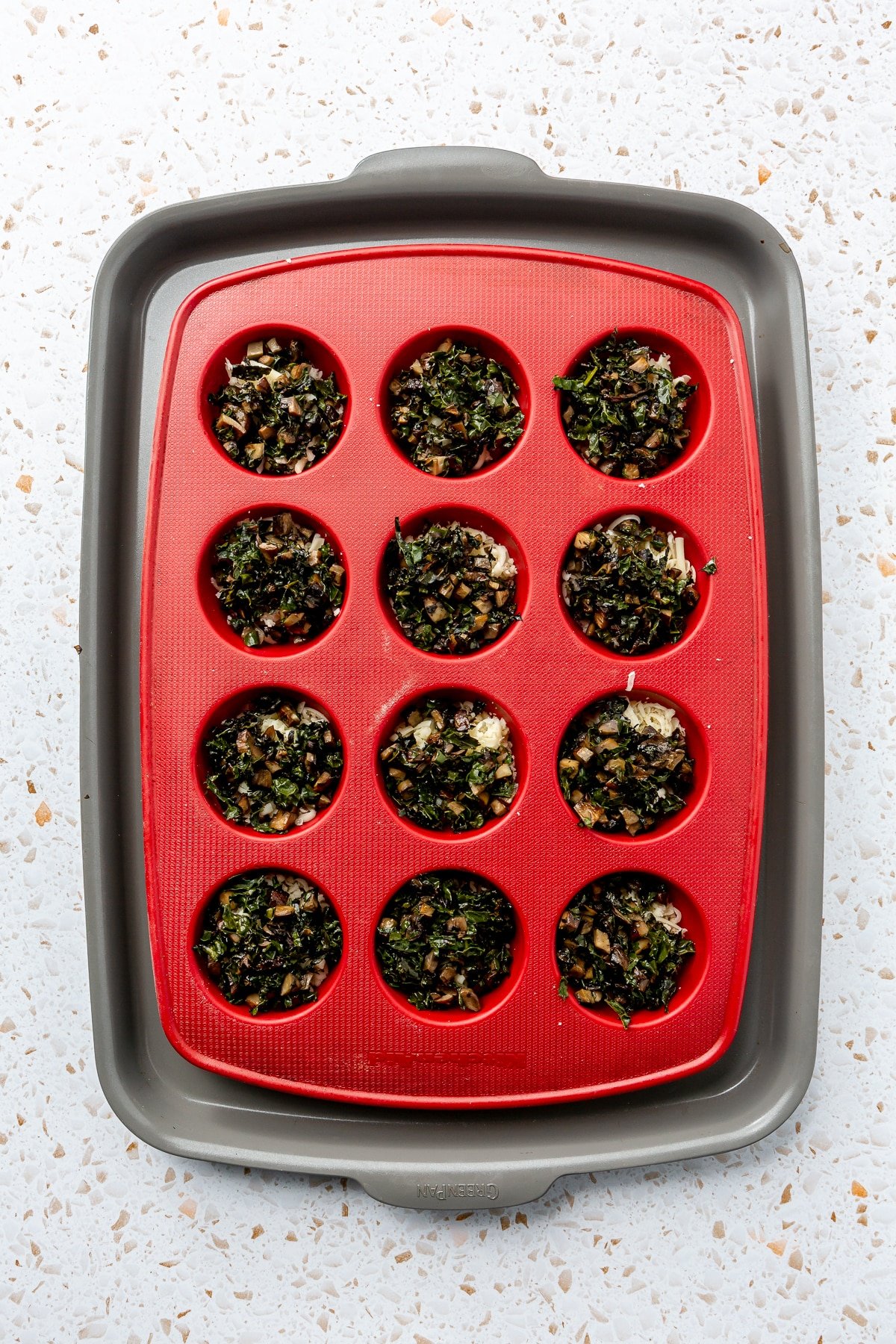 The roasted mushrooms and kale have been placed into a red, silicone muffin tray which sits on a metal baking tray.