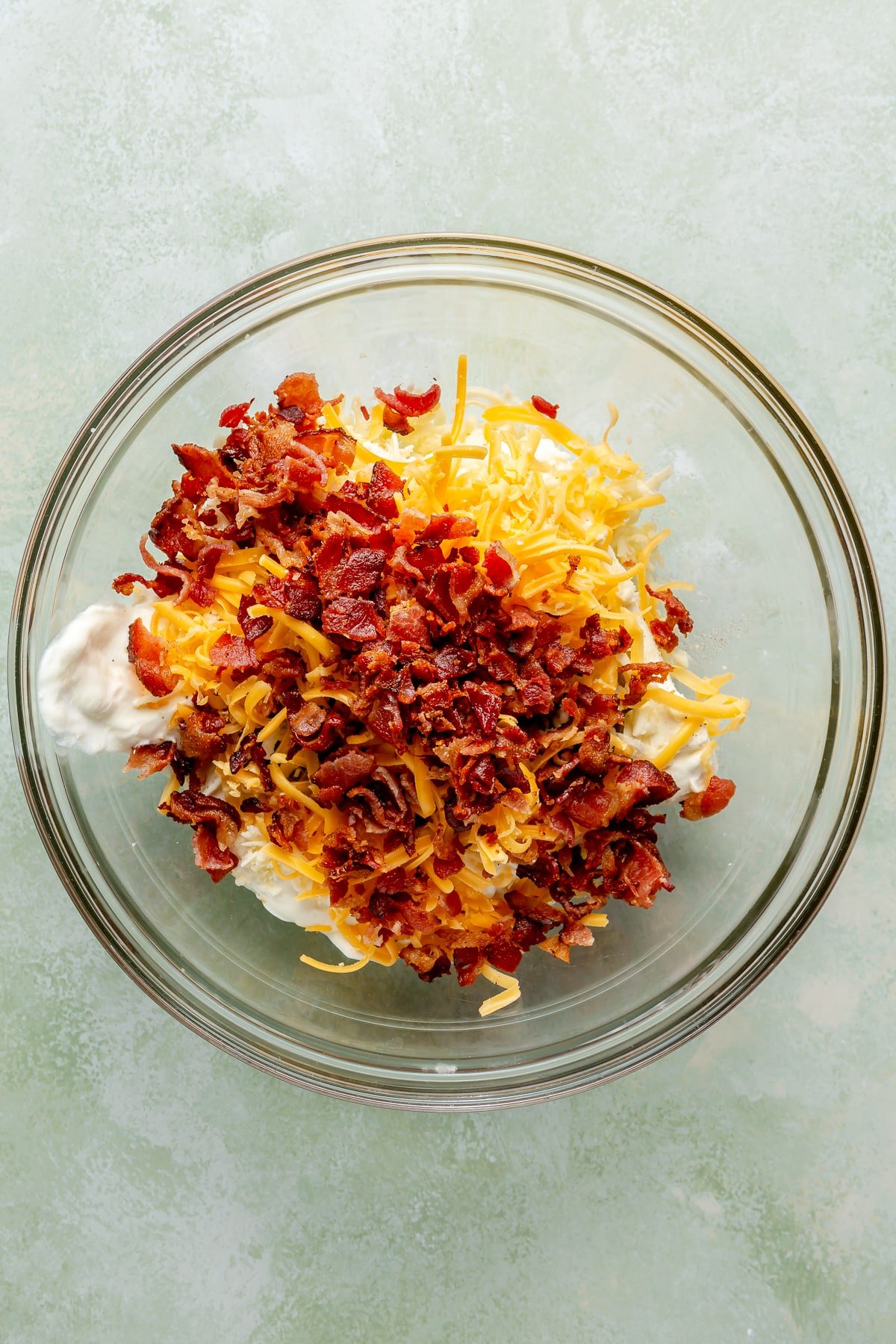 Cream cheese, shredded cheddar, and bacon pieces have been placed into a glass mixing bowl.